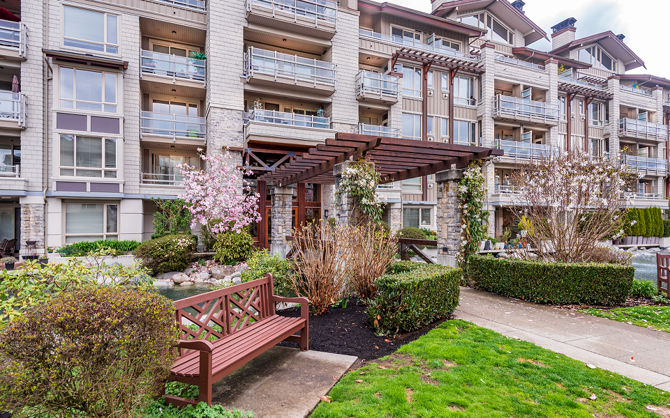 Modern senior living residence with glass balconies, landscaped gardens, a wood bench, and flowering trees.