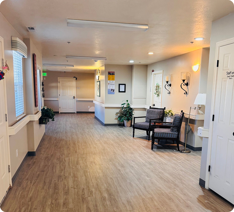 Bright senior living hallway with wood floors, handrails, and a seating alcove with patterned armchairs.
