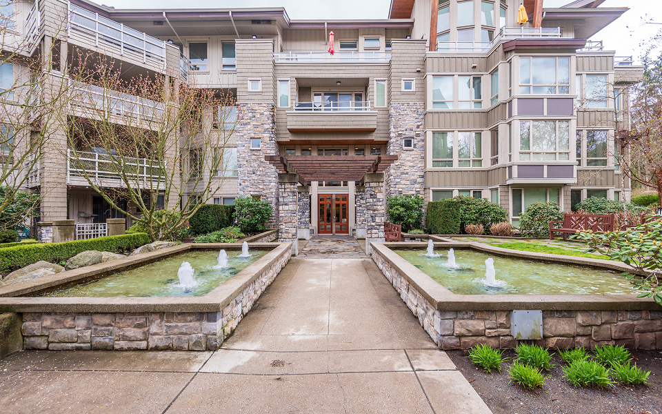 Entrance to a multi-story senior living community with stone fountains lining a paved walkway.